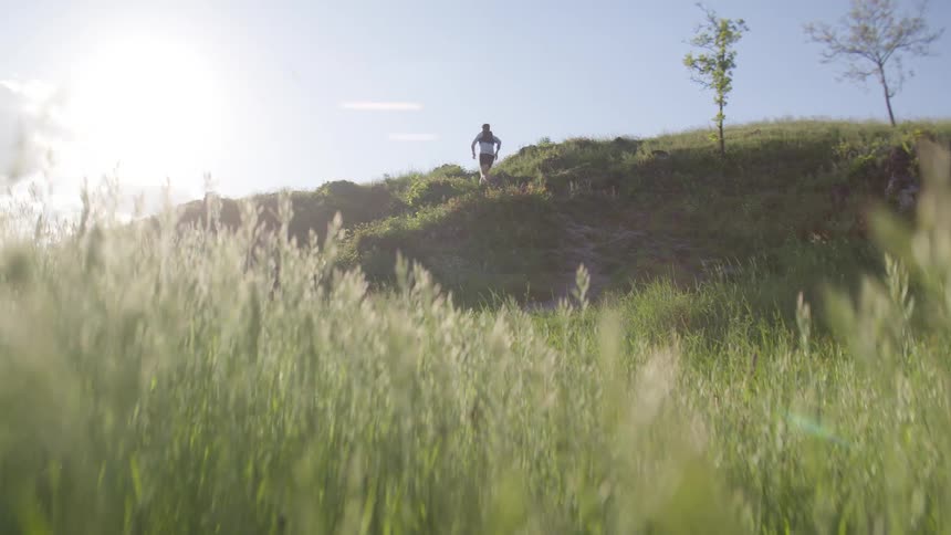 Man runs uphill with tall grass in the foreground and a clear sky above