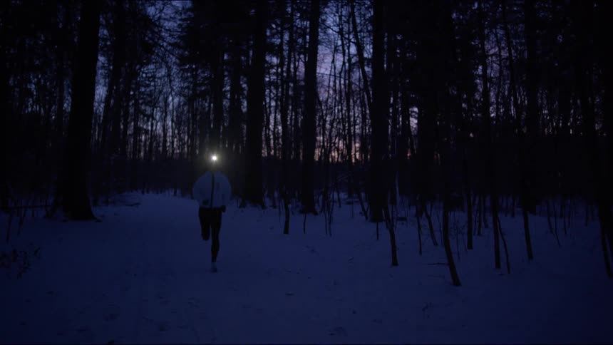 A woman running across a snowy forest at night with a lantern attached to her head