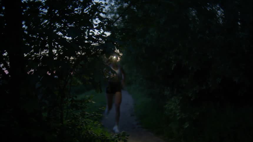 Woman with a flashlight on her head running along a trail with trees around on a dark morning