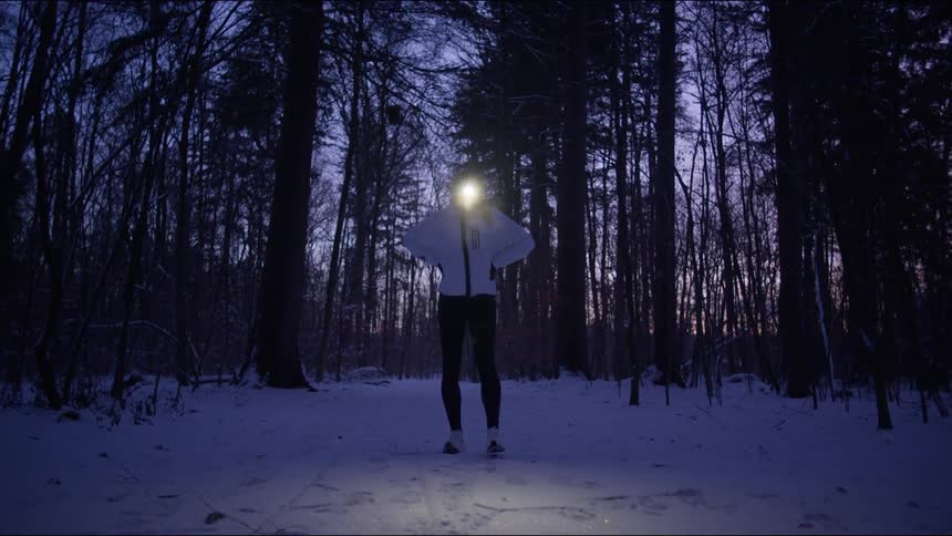 Woman standing in a snowy conifer forest wears a bright headlamp and stretches