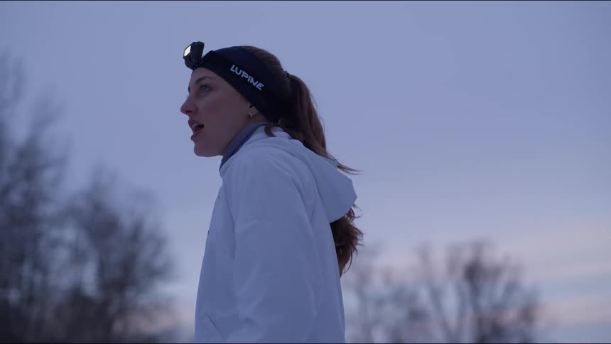 Woman wearing a headlamp stands between groves of leafless trees and looks around
