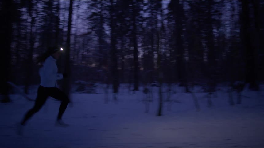A runner illuminated by a headlamp jogging through a dark snowy forest