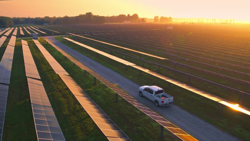 A truck drives on a road between solar panel rows as the sun shines bright on the horizon