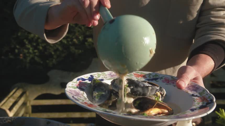 Man ladles shellfish in a white sauce into a large bowl while standing outside