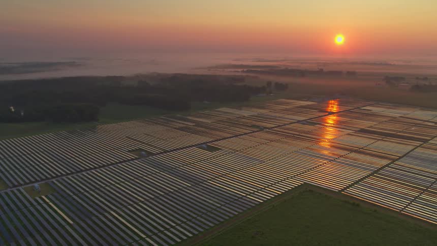 Large field of solar panels at dawn with the sun rising over the horizon and reflecting off the panels