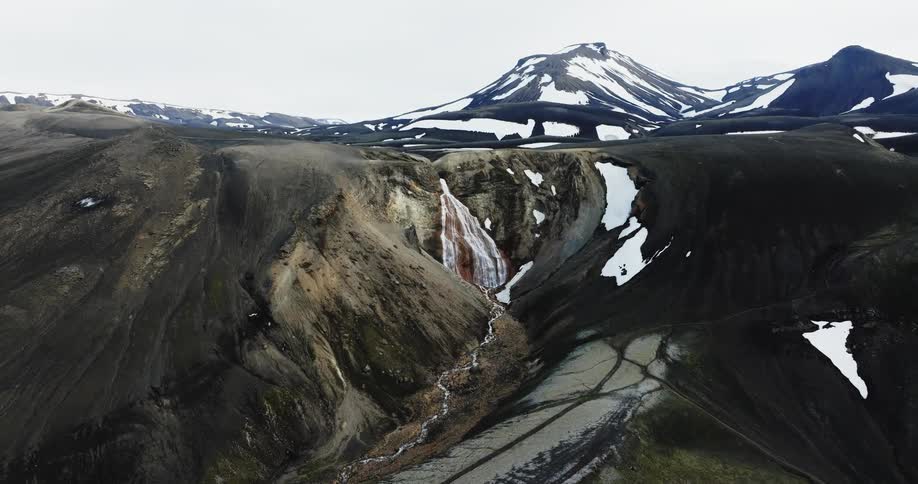 A small waterfall coming out of the peak of a stone hill in a mountainous region with some snow on the mountains