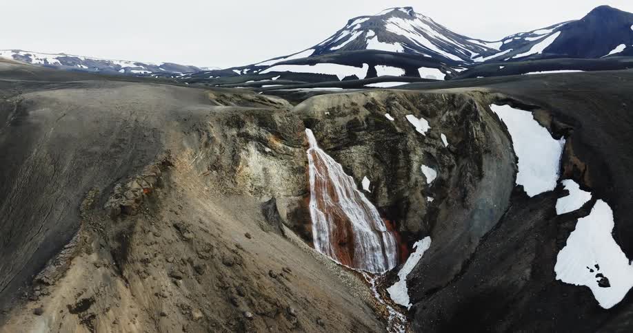A small waterfall coming out of rocks from a column in a mountainous region with some snow on them