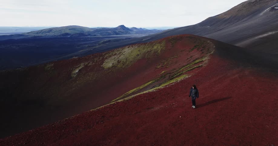 A person walking along the crest of a red volcanic crater set against a vast mountainous landscape