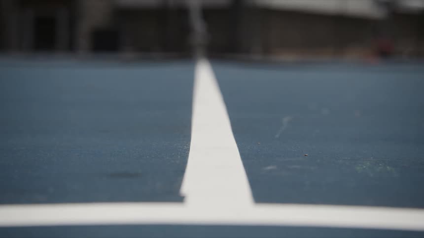 A tennis ball rolling on the floor of a blue court with a white line in the center and the ball going left and right
