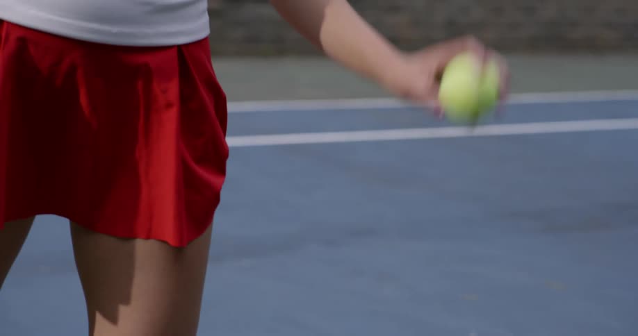 A female tennis player in a red skirt bounces a tennis ball on a court