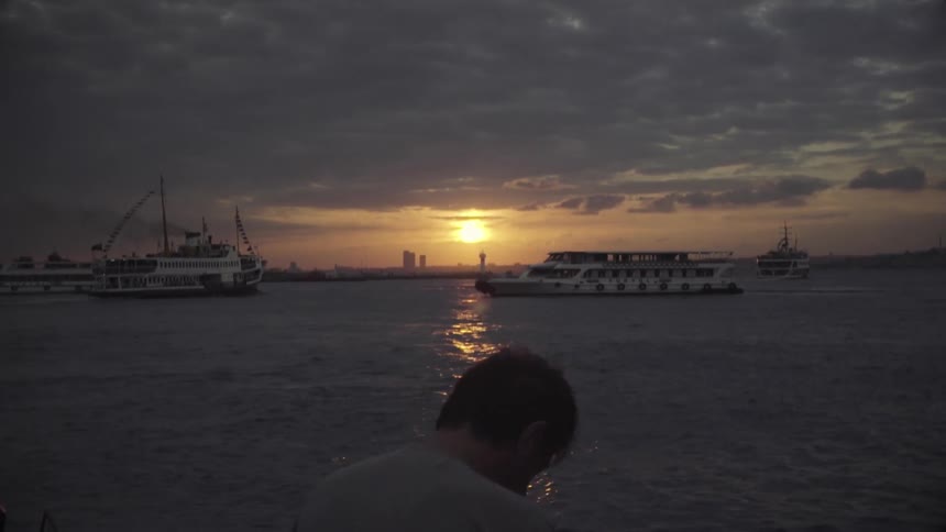 A man standing near the ocean with boats coming in at sunset