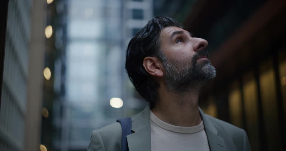 An older man with a beard looks around at tall city buildings in a downtown area