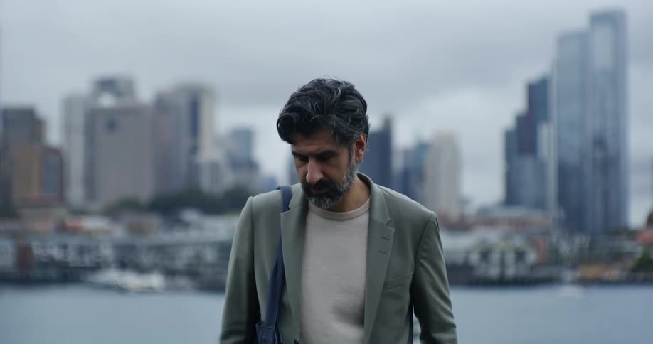 A bearded man in formal clothes stands and adjusts his suit jacket and then stares straight ahead