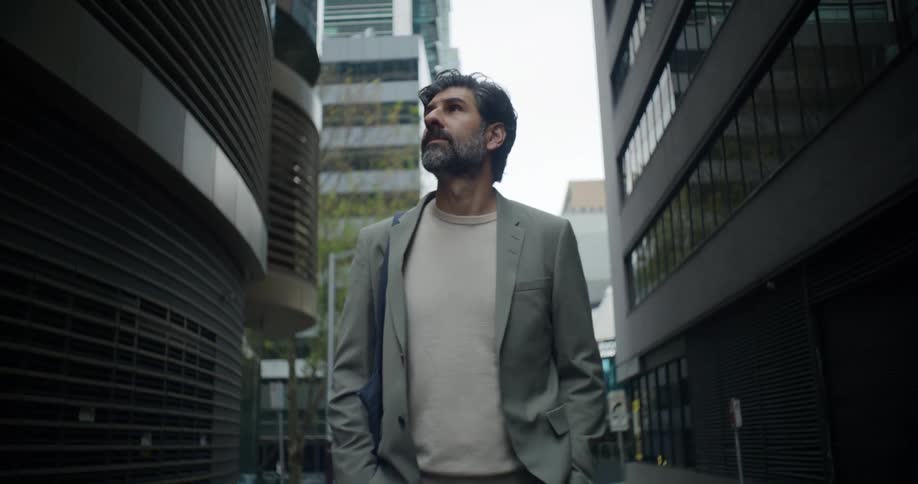 Man in a gray suit with a crossbody bag and a beard walking down a street while looking at the large modern buildings
