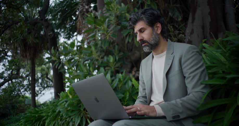 Man in gray suit sitting in natural environment with greenery typing on laptop