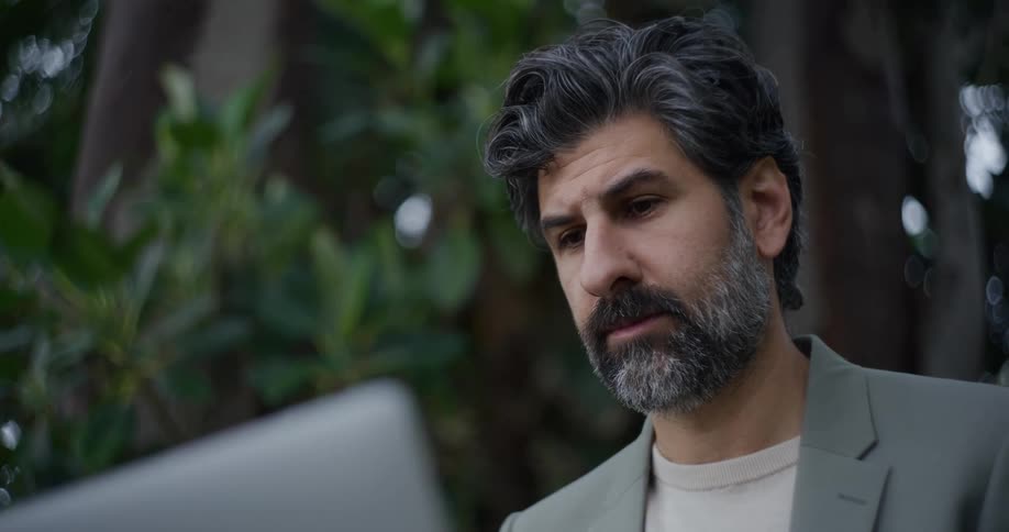 Bearded man wearing a gray blazer sitting using a laptop in an outdoor setting