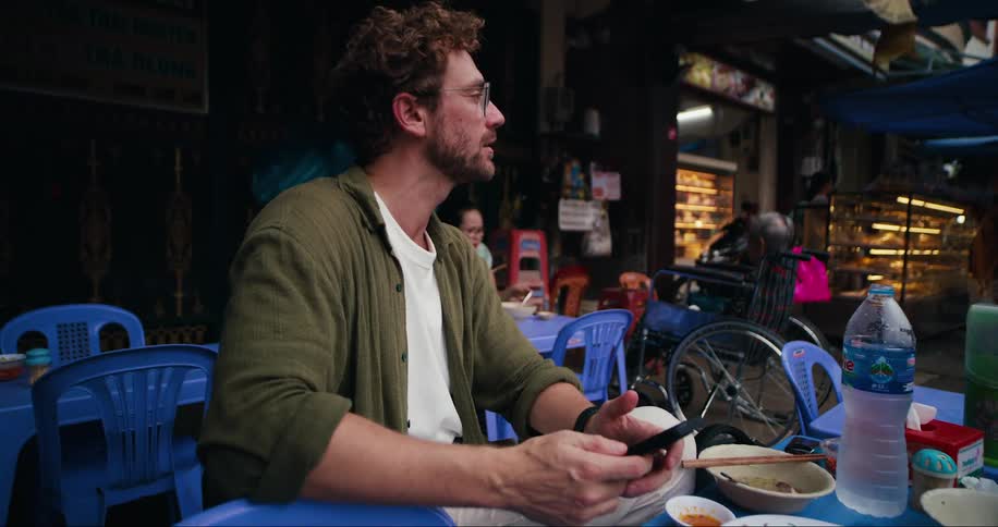 A young man sits at an outdoor restaurant after finishing a meal texting on his phone
