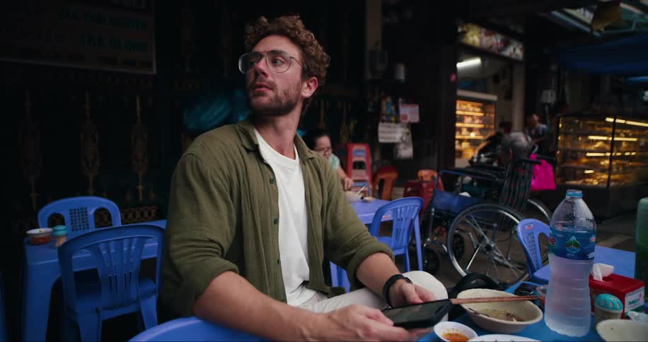 A man wearing glasses is sitting on a blue chair and has a cell phone in his hand and he looks to his right side looking for something