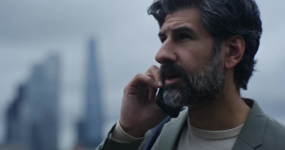 A bearded man talking on a smartphone with a city skyline in the background