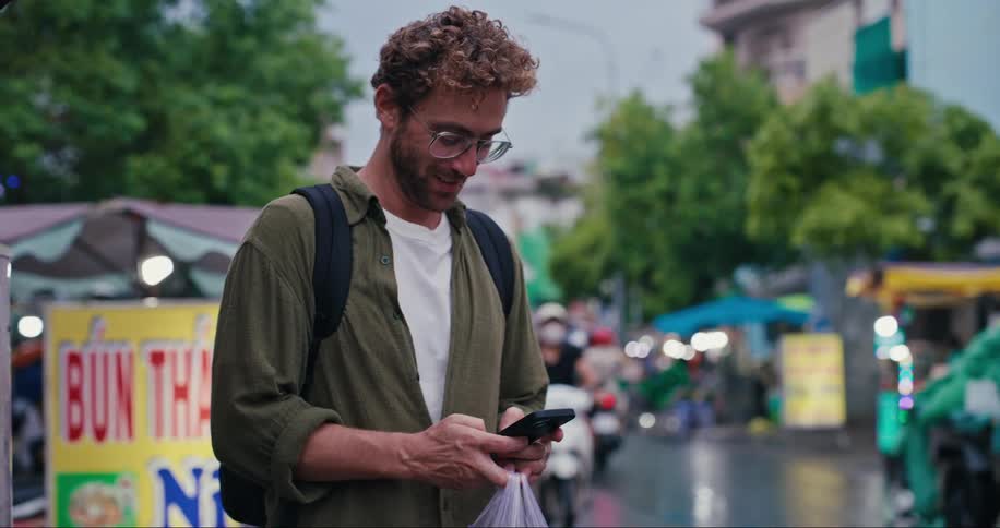 A man is standing by a road where multiples scooters go by while smiling and writing on his smart phone