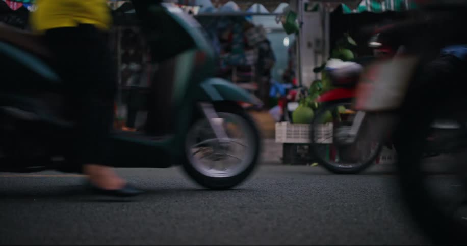 An asphalt street with several motorcycles passing by and on the sidewalk there is a small fruit market