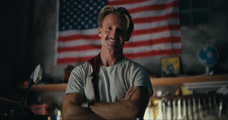 A man slaps a rag across his shoulder and smiles while standing next to a motorcycle in a garage