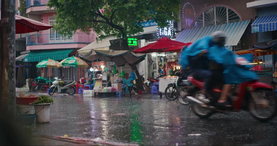 Several people driving motorcycles on an asphalt street and it is raining