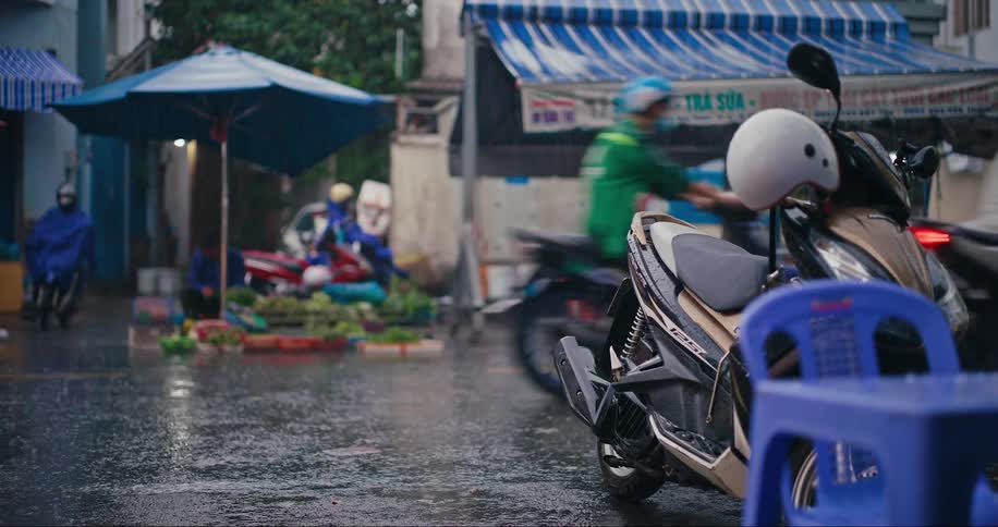 A scooter parked in a street while it rains with multiple other scooters passing by behind it