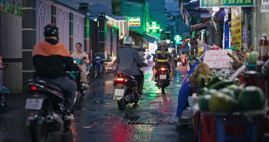 Narrow wet street with motorcyclist traffic and small markets selling food and fruit all around