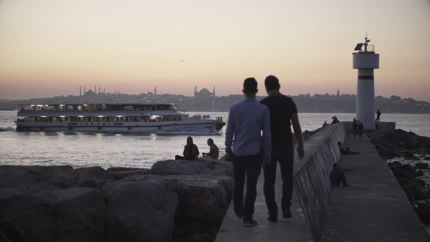 Two men walking on concrete pier towards lighthouse and ocean bay