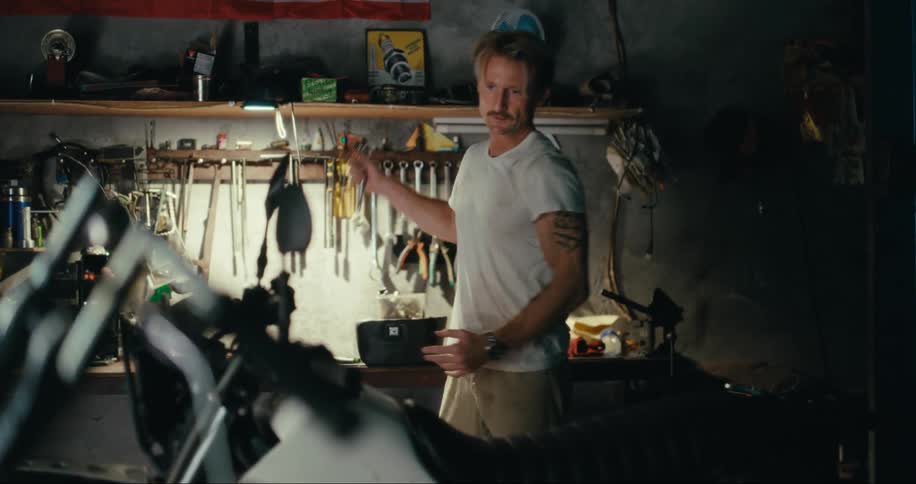 A man walks into a garage carrying a box from which he takes out a tool that he uses on a motorcycle