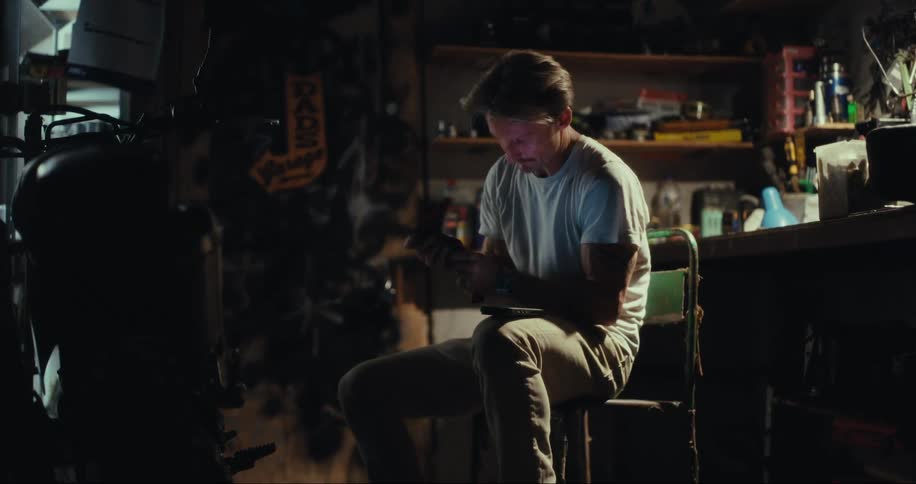 A man sitting in a dark workshop makes a phone call while using a rag to clean his hands