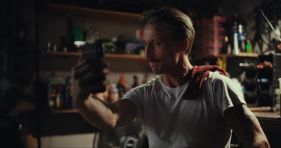 A blond man with a mustache is looking at a cell phone and he is laughing and then he runs his hand over his chin