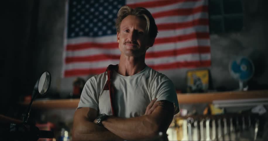 A white man throws a dirty rag over his shoulder while standing by a motorcycle and in front of an american flag in a garage