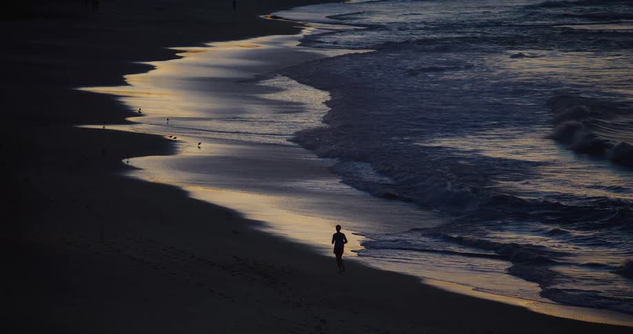 Silhouette of a jogger running on a sandy shoreline as the waves gently roll in