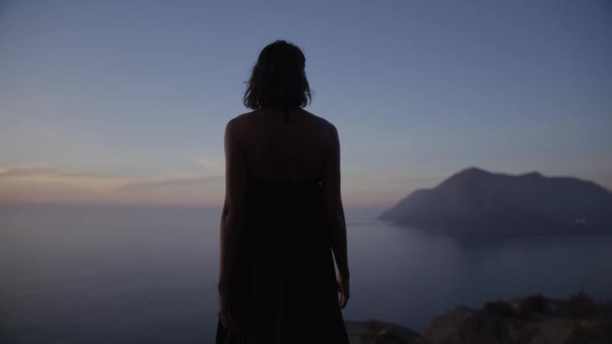 Back view of a woman in a long dress walking on a cliff by the sea at dawn
