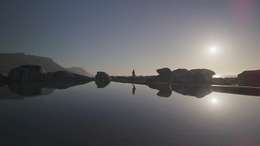 A woman walks near a lake surrounded by large boulders with the sun shining on a clear morning sky