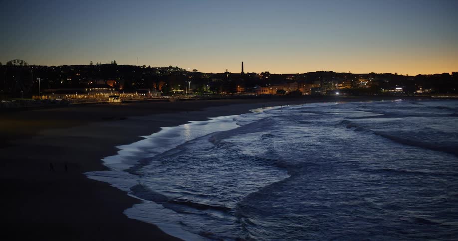 Beach at dusk with gentle waves lapping the sand and city lights in the background illuminating the skyline