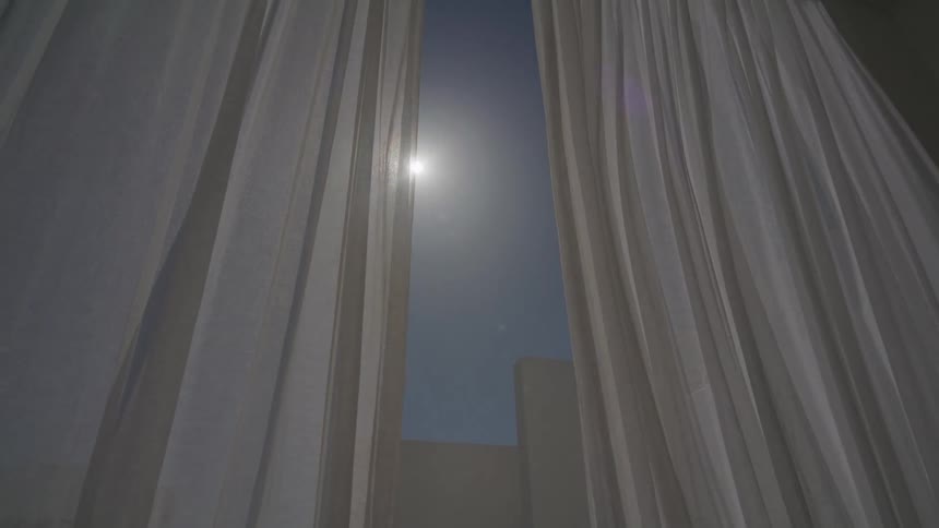 Some white curtains moving due to the wind next to a glass window overlooking the blue sky and the sun shining