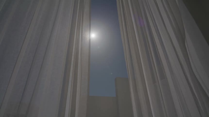 White curtain moving through the air on a glass door overlooking a clear blue sky and the sun in the spotlight