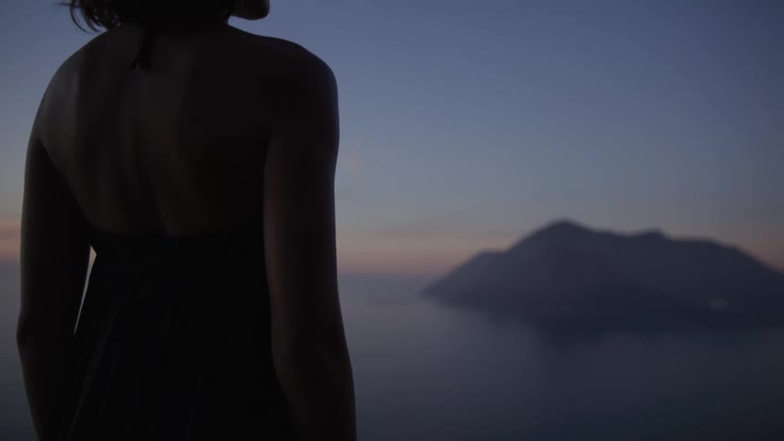A woman stands next to a body of water with mountains in the distance at dusk