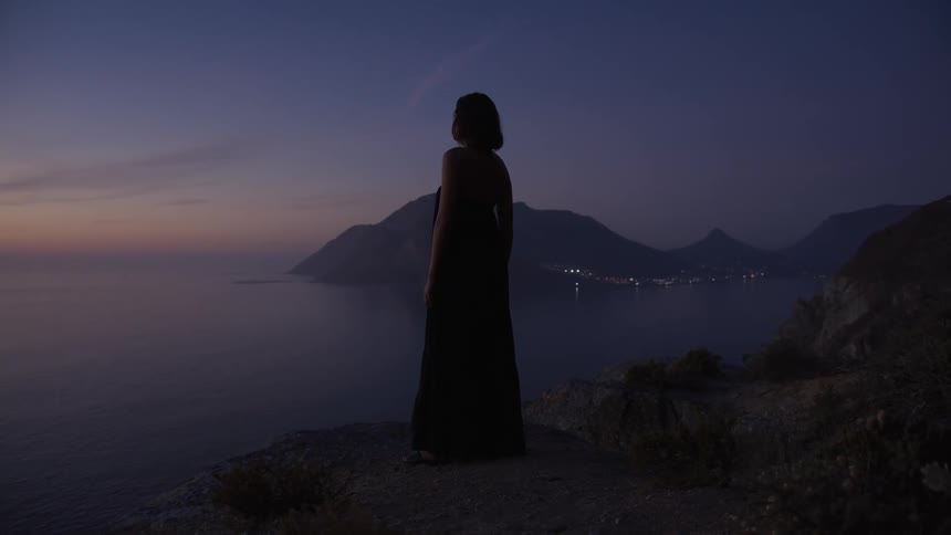 Woman in long dress on a cliff by the sea on a day with purple and orange sky