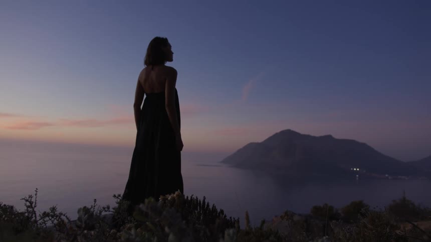 A woman in a dress is standing with her back turned and walking to a shore to look at an island in the middle of an ocean on a morning