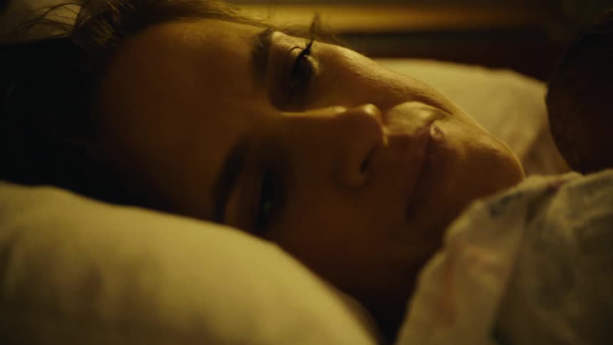New mother lying in bed with her newborn sleeping on her chest turns her face and grimaces