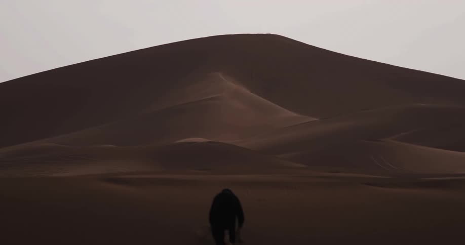 A man wearing a black robe is climbing a sand dune in the desert