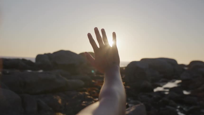 Womans hand towards the sun and its reflection in a natural environment with rocks by the sea