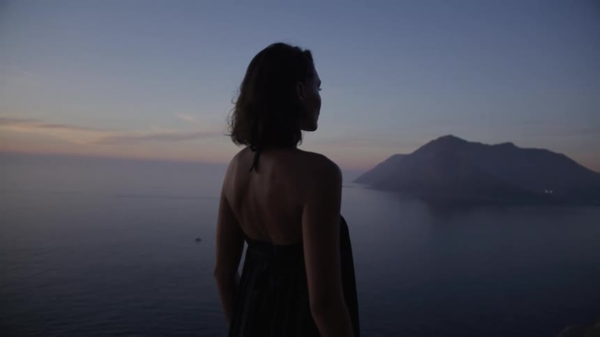 Woman from behind contemplates a serene landscape of mountains and sea during twilight