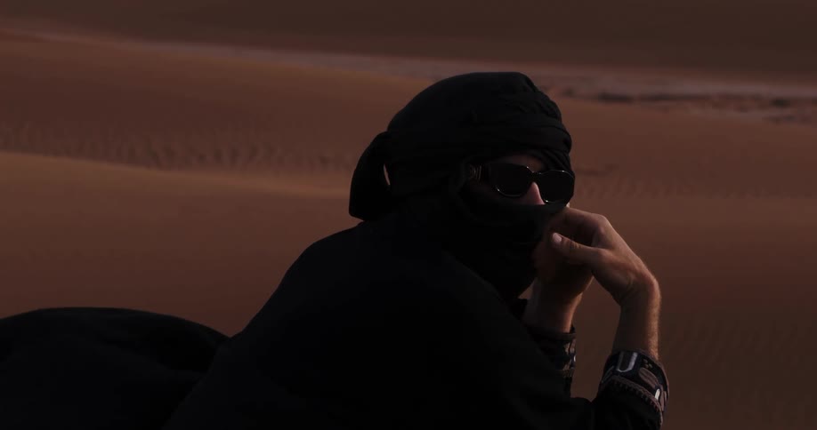 Man with black clothing covering his face standing in the middle of dunes in the desert