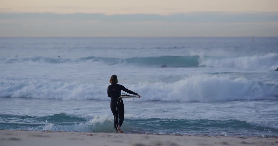 Female sufer carrying a surfboard walking in to the ocean towards the waves