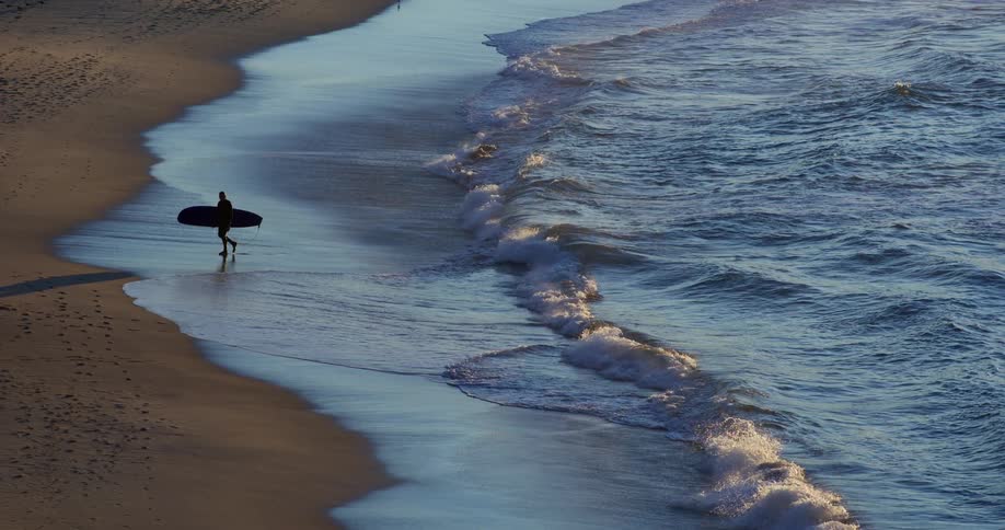 Silhouette of a person with a long surfboard coming out of the sea and walking on the sand of the beach